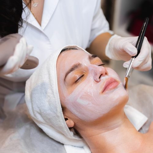 Beautician with a brush applies a white moisturizing mask to the face of a young girl client in a spa beauty salon